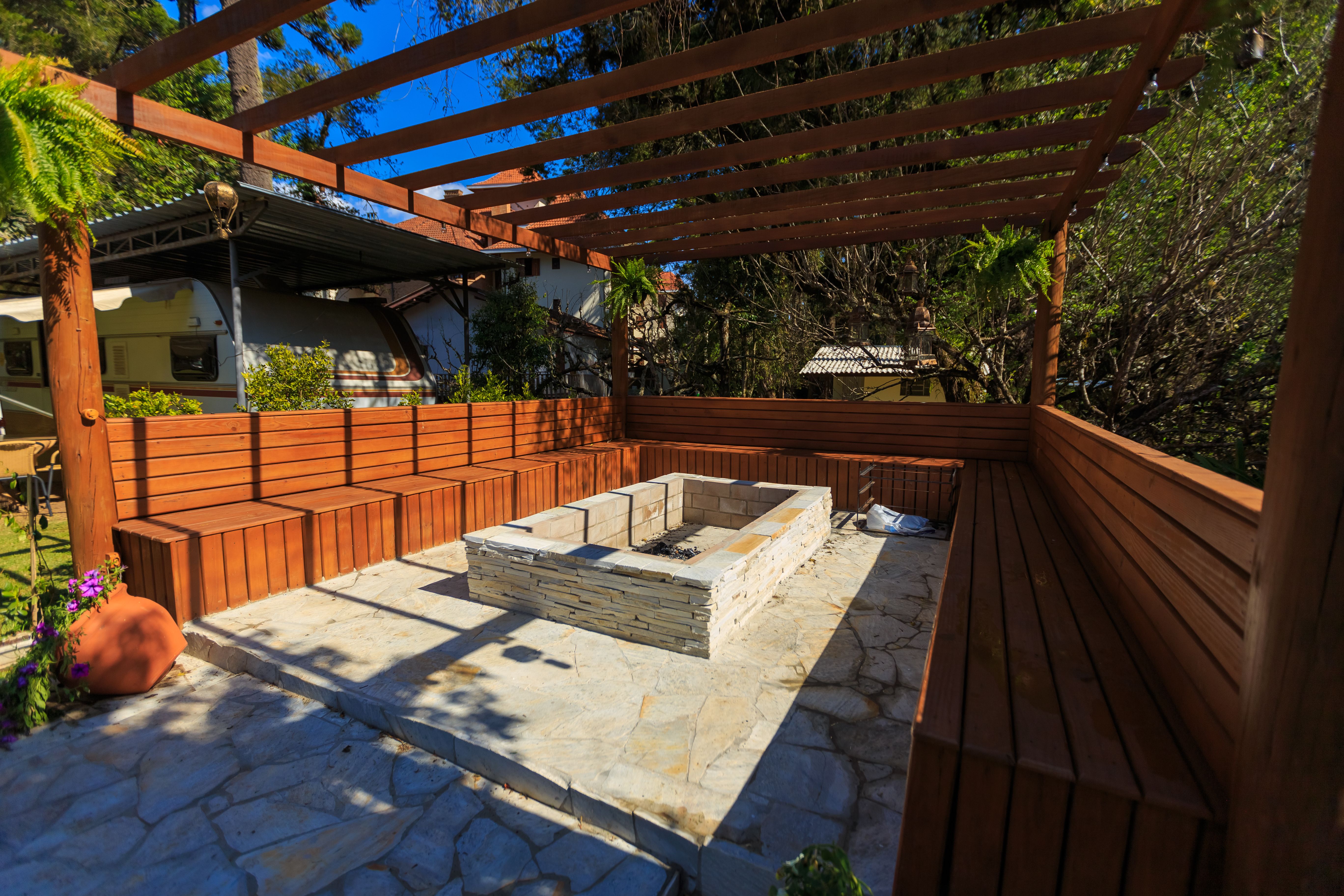 Outdoor seating with a stone fire pit beneath a pergola in a landscaped backyard.